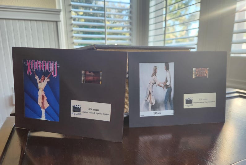 May include: Two black display cases with film strips and movie posters. One case features the word "XANADU" in pink and a woman in a white outfit. The other case has a photo of two people and the word "XANADU". Both cases have a label that says "35 mm Original Insert Special Edition".
