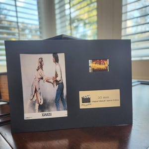 May include: A black framed display featuring a film strip and two images from the movie "Xanadu." The larger image shows two people holding hands. The text "XANADU" is visible. A smaller image and a label that reads "35 mm Original Minicell Special Edition" are also present.