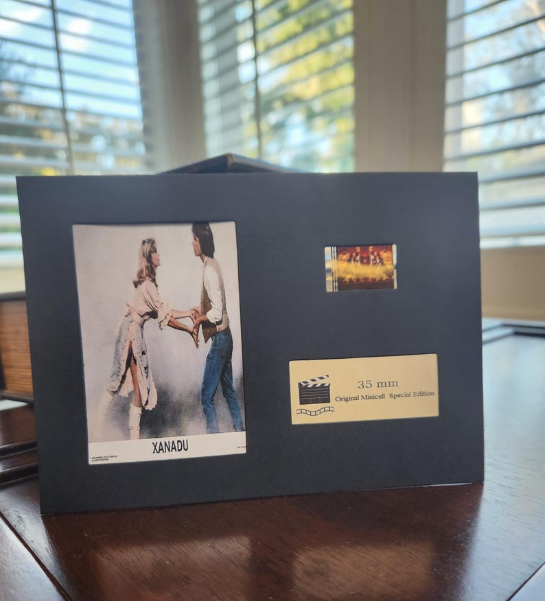 May include: A black framed display featuring a film strip and two images from the movie "Xanadu." The larger image shows two people holding hands. The text "XANADU" is visible. A smaller image and a label that reads "35 mm Original Minicell Special Edition" are also present.