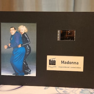 May include: A framed print featuring a photograph of two people, one in a blue outfit and the other in a sequined black outfit. The frame also includes a small film strip and a label that reads "Madonna".