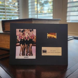 May include: A black framed display featuring a vintage photo of women in pink tops and black skirts, with the word "XANADU". Includes a film strip and a label that reads "35 mm Original Moviecell Special Edition".
