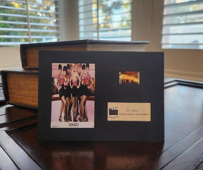 May include: A black framed display featuring a vintage photo of women in pink tops and black skirts, with the word "XANADU". Includes a film strip and a label that reads "35 mm Original Moviecell Special Edition".