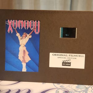 May include: A brown filmcell display featuring the "Xanadu" movie poster with a woman in white. It includes a film strip and a label stating "Original Filmcell Special Edition 35 MM."