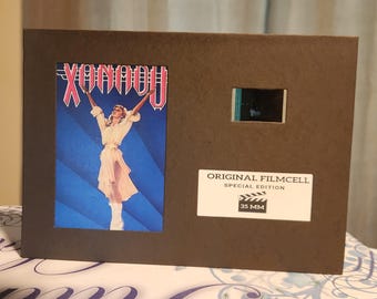 Op de afbeelding: Een bruine filmcel-display met de filmposter van "Xanadu" met een vrouw in het wit. Het bevat een filmstrip en een label met de tekst "Original Filmcell Special Edition 35 MM."