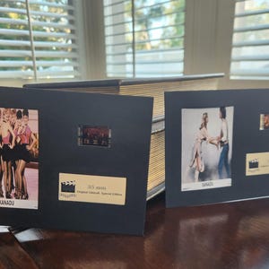 May include: Two black framed film cells from the movie Xanadu. One frame features a group of women in pink tops and black skirts. The other frame shows a couple dancing. Both frames have a small film strip and a gold label.