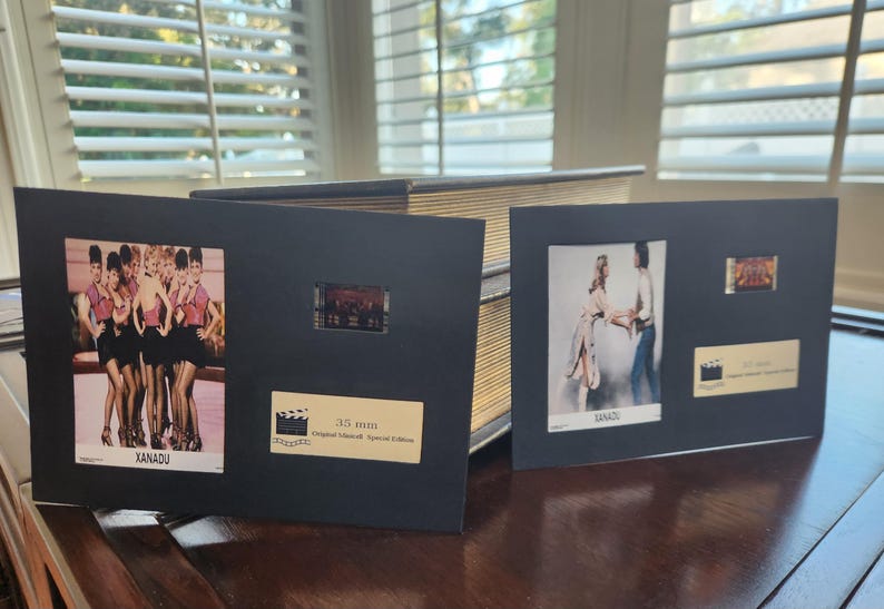 May include: Two black framed film cells from the movie Xanadu. One frame features a group of women in pink tops and black skirts. The other frame shows a couple dancing. Both frames have a small film strip and a gold label.