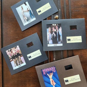 May include: Four framed film cells from the film "Xanadu" are displayed on a wooden surface. Each frame features a small image from the film, a film strip, and a label. The frames are in shades of grey and brown.