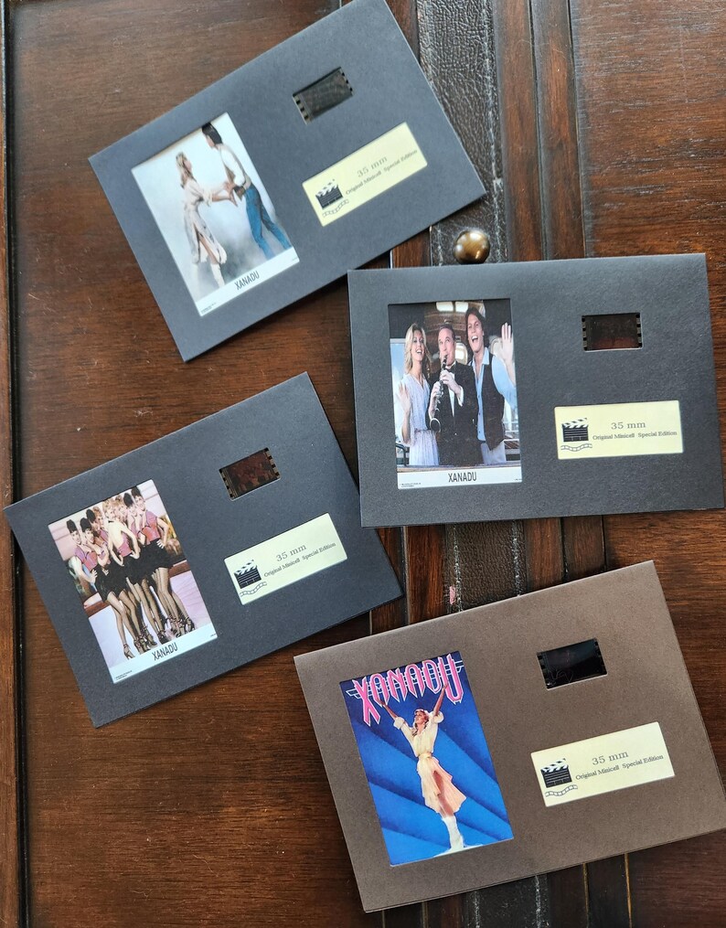 May include: Four framed film cells from the film "Xanadu" are displayed on a wooden surface. Each frame features a small image from the film, a film strip, and a label. The frames are in shades of grey and brown.