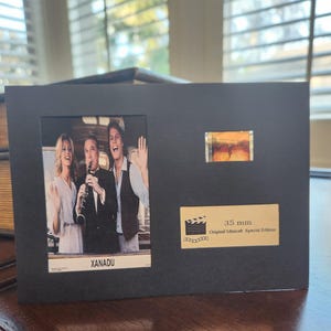 May include: Black framed print featuring a still from the movie "Xanadu" with three actors. Includes a small film strip and a label that reads "35 mm Original Minicell Special Edition".