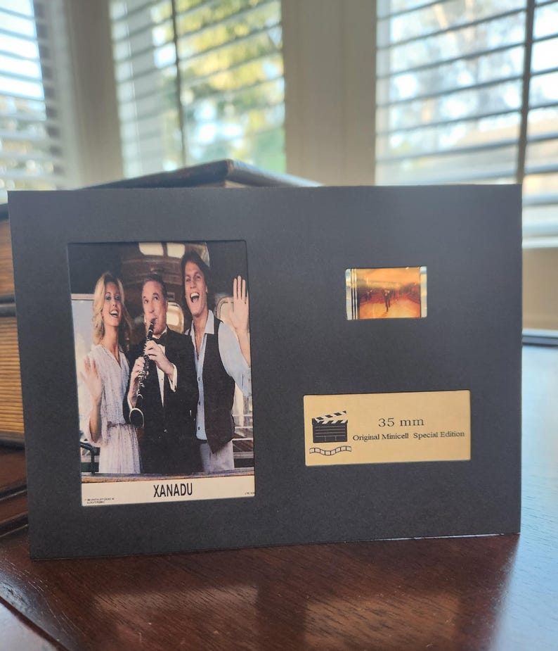 May include: Black framed print featuring a still from the movie "Xanadu" with three actors. Includes a small film strip and a label that reads "35 mm Original Minicell Special Edition".