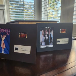 May include: Two framed film cells, one with a movie poster for "Xanadu" and the other with a still from the film "Xanadu". Each frame includes a film strip and a label with text, presented on a dark surface.