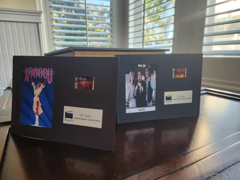 May include: Two framed film cells, one with a movie poster for "Xanadu" and the other with a still from the film "Xanadu". Each frame includes a film strip and a label with text, presented on a dark surface.