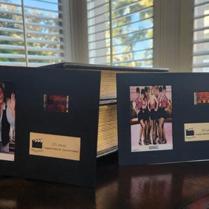 May include: Black framed movie memorabilia featuring stills and film strips from the film Xanadu. The frames include images of the cast and the film's logo. The frames are displayed on a dark wooden surface.