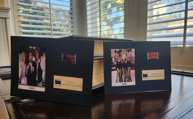 May include: Black framed movie memorabilia featuring stills and film strips from the film Xanadu. The frames include images of the cast and the film's logo. The frames are displayed on a dark wooden surface.