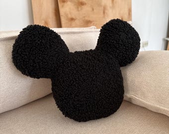 May include: Black plush pillow shaped like Mickey Mouse's head with large ears. The pillow is sitting on a light beige couch.