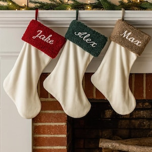 May include: Three personalized Christmas stockings hang above a brick fireplace. Each stocking has a cream-colored body and a textured cuff in red, green, and brown. Names Jake, Alex, and Max are embroidered on the cuffs.
