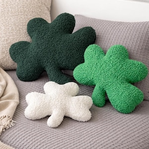 Peut inclure: Quatre coussins décoratifs en forme de trèfle, dans des tons de vert et de blanc, reposent sur un lit. Les coussins ont un aspect texturé et moelleux. L'arrière-plan comprend des coussins aux tons neutres et un couvre-lit gris.