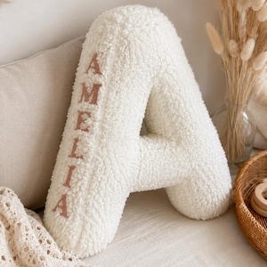 Puede incluir: Un gran cojín blanco y esponjoso con forma de letra "A", con el nombre "AMELIA" bordado en rosa suave. El cojín tiene un aspecto texturizado y afelpado y está colocado sobre una superficie de tonos neutros, lo que sugiere una habitación infantil.