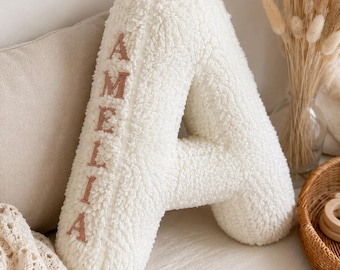 May include: A large, fluffy white letter "A" pillow with the name "AMELIA" embroidered in a soft pink. The pillow has a textured, plush appearance and is placed on a neutral-toned surface, suggesting a nursery or children's room.