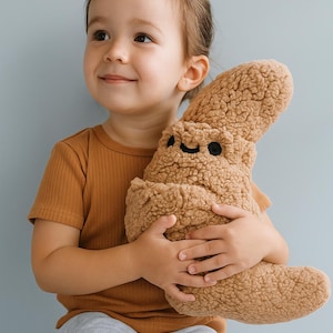 May include: A plush, brown stuffed animal with a smiling face is held by a child. The toy has a textured, fuzzy appearance. The child is wearing a brown ribbed t-shirt and gray sweatpants. The background is a solid light blue.