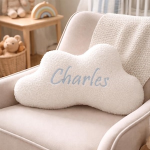 May include: A cloud-shaped decorative cushion made of white textured sherpa fabric. The centre of the cushion features the name Charles embroidered in light blue script lettering. The cushion has a soft, plush appearance with rounded edges.