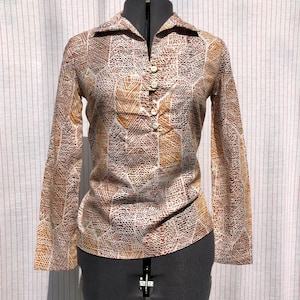 May include: A long-sleeved blouse with a brown, tan, and white geometric pattern. The blouse has a collared neckline and a button-down front. The fabric appears to be lightweight, and the blouse is displayed on a mannequin.