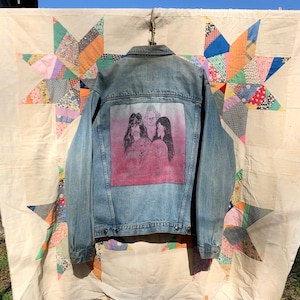 May include: A light blue denim jacket with a faded pink graphic on the back featuring three figures. The jacket is hanging on a clothesline in front of a colorful star-patterned quilt.
