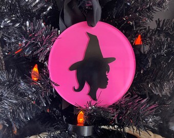 May include: A pink round ornament with a black silhouette of a witch wearing a hat. The ornament is hanging on a black artificial Christmas tree.