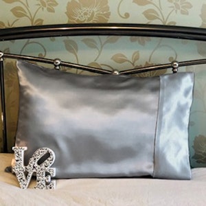 May include: A silver satin pillowcase with a contrasting border. The pillowcase is rectangular and sits on a bed with a floral patterned headboard. A decorative "LOVE" sign is in the foreground.