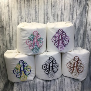 May include: Five white toilet paper roll covers with embroidered decorative swirls and the letter "A" in different colors. The colors are pink, purple, blue, black, and brown.