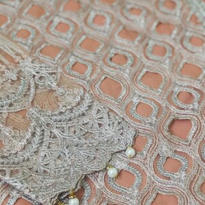 May include: A close-up of a peach-colored fabric with a silver embroidered pattern. The fabric has a delicate, lacy texture and is adorned with small, white pearl-like embellishments.
