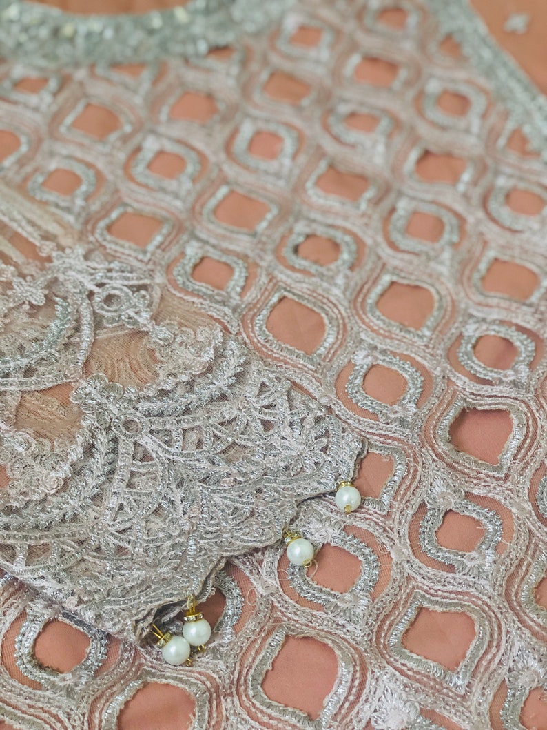 May include: A close-up of a peach-colored fabric with a silver embroidered pattern. The fabric has a delicate, lacy texture and is adorned with small, white pearl-like embellishments.