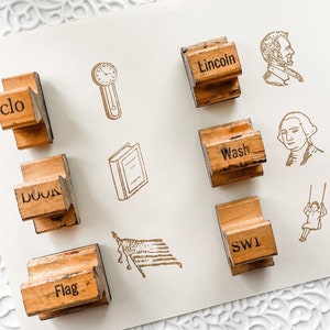 Puede incluir: Un conjunto de sellos de madera con varias palabras e imágenes, incluyendo "clo", "book", "Flag", "Lincoln", un reloj, un libro y retratos. Los sellos están dispuestos sobre una superficie blanca.