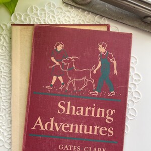 May include: A vintage book titled "Sharing Adventures" with a maroon cover. The cover features illustrations of two children and a goat. The book is resting on a white lace doily, with a stapler and plant in the background.
