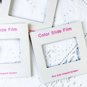 Könnte beinhalten: Mehrere graue Diaschutzhüllen mit dem pinkfarbenen Text "Color Slide Film". Die Dias liegen auf einer weißen, strukturierten Oberfläche mit einem spitzenartigen Muster. Der Text "this side toward screen" ist ebenfalls sichtbar.