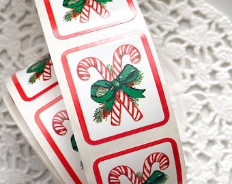 May include: A roll of square Christmas stickers. Each sticker features a red-and-white candy cane design with a green bow and sprigs of greenery, set against a white background with a red border. The stickers are on a roll.