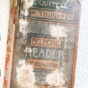 May include: A close-up of an antique book titled "McGuffey's Third Eclectic Reader, Revised Edition." The cover is brown with black and gold lettering and decorative scrollwork. The book shows signs of age and wear.