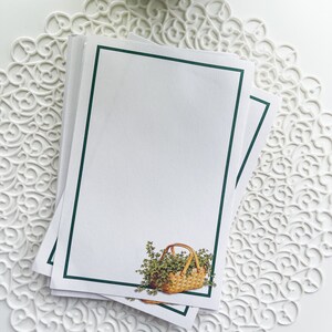 May include: A stack of blank note cards with a green border. The top card features a small illustration of a wicker basket filled with green leafy plants.