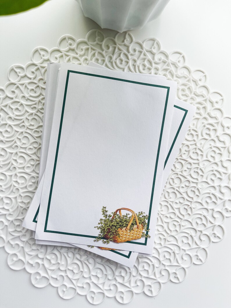 May include: A stack of blank note cards with a green border. The top card features a small illustration of a wicker basket filled with green leafy plants.