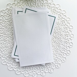May include: Stack of white cards with a dark green border on a white doily.