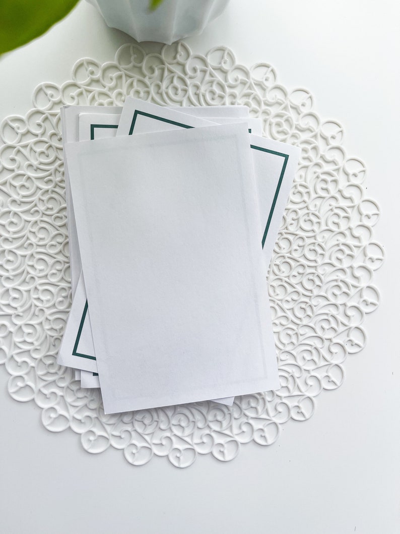 May include: Stack of white cards with a dark green border on a white doily.