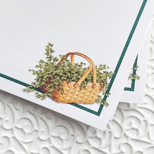 May include: A white card with a green border and a basket filled with green leaves. The basket is made of woven wood and has a brown handle.