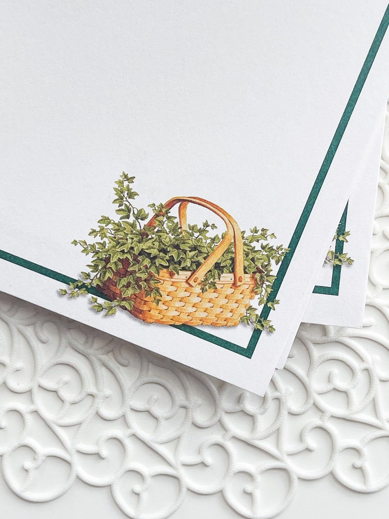 May include: A white card with a green border and a basket filled with green leaves. The basket is made of woven wood and has a brown handle.