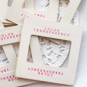 May include: A collection of vintage color transparency slides with the text "COLOR TRANSPARENCY" in pink. The slides are cream-colored and have a rectangular window revealing a white, lacy doily. The slides are scattered on a white surface.