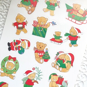 Op de afbeelding: Een vel stickers met verschillende kerstthema-illustraties van beren. De beren dragen feestelijke kleding en houden items vast zoals cadeaus, snoepstokken en kransen. Sommige beren zijn afgebeeld met hun jongen.