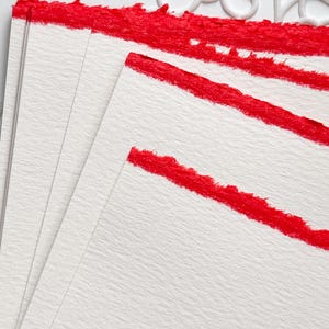 Puede incluir: Una pila de hojas de papel rectangulares blancas con bordes texturizados y desiguales. Cada hoja presenta una banda horizontal gruesa de pigmento rojo vibrante aplicada en el borde superior, creando un contraste audaz con la superficie blanca.