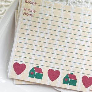 Könnte beinhalten: Ein Stapel Rezeptkarten mit cremefarbenem Hintergrund und Karomuster. Die Karten haben Linien zum Schreiben und zeigen rote Herzen sowie grüne und rote Hausillustrationen. Auf der Oberseite der Karten steht "Recipe:" und "Recipe From:"