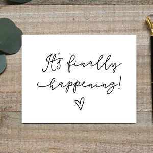 May include: A white card with the text "It's finally happening!" written in black cursive with a small heart drawn below the text. A gold-colored pen with a black ink cartridge is lying on a wooden surface next to the card. A sprig of eucalyptus is in the upper left corner of the image.
