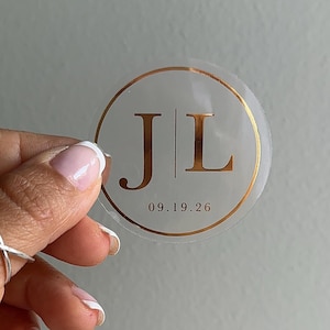 May include: Clear round sticker with gold foil accents. The sticker features the initials "JL" in a serif font, separated by a vertical line, and the date "09.19.26" in a smaller font below. The sticker is held up against a gray background.