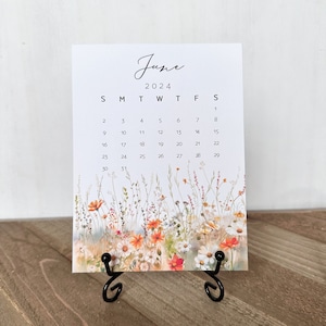 May include: A June 2024 calendar with a floral design featuring white, orange, and red flowers. The calendar is on a black metal stand.